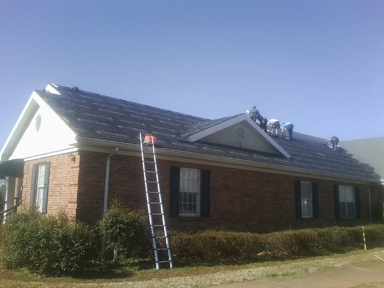 Crew tearing shingles off a brick ranch roof with a ladder set — Emergency Roof Repair work by our crew in Milner