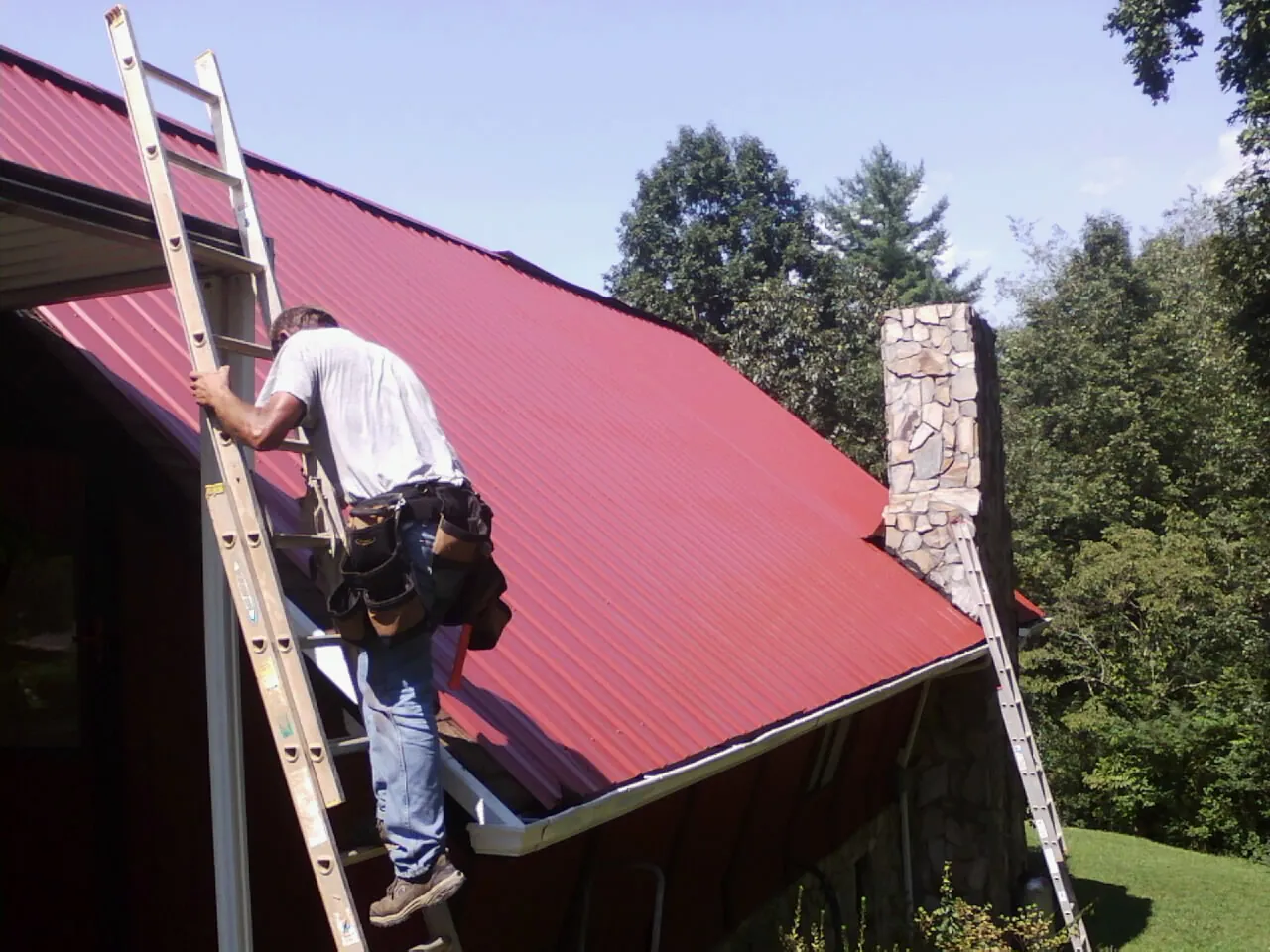 Roofer on a ladder beside a red metal roof and stone chimney — Soffit & Fascia Repair by Roofing Brothers in Milner